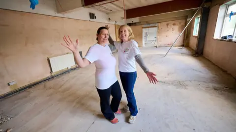 Two women wearing white t shirts and dark coloured trousers stand in a room which has bare plaster walls and floorboards, with their arms outstretched. 