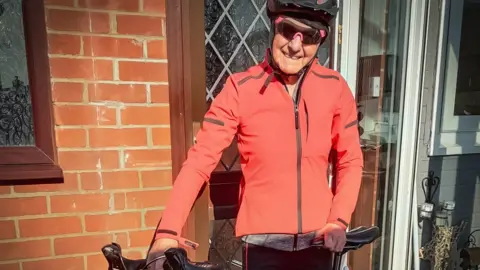 Family photo Harold standing in front of the glass door entrance to brick home, holding a racing bicycle and looking fit in biking clothing: jacket, glasses and safety helmet