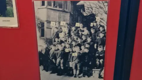 A black and white picture showing children standing outside Hawes End during the war. They are all wearing shorts and jackets and are standing in a large group formation, with some adults at the back. The photo is grainy and is affixed on a red noticeboard.