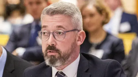 Liam McBurney/PA Wire Niall Ó Donnghaile pictured in October 2022. He is wearing thick-rimmed glasses, and he has a greying beard and grey hair. He is wearing a dark suit, a white shirt and a dark striped tie