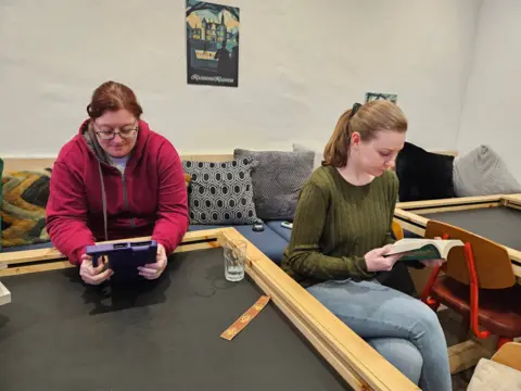 Two readers of the silent book club read while sitting around a board game table. One, with reddish-brown hair, is reading from a purple tablet. The other, with lighter brown hair, is reading a paperback book.