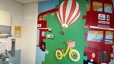 A mural painted on a hospital wall depicting a red double decker bus with green jigsaw pieces on top of it showing an air balloon and a child's bike.