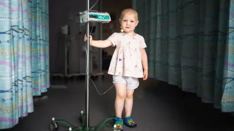 Kris Askey / British Journal of Photography A photo of a little girl standing in the camera, she is wearing shorts and a top and holding onto a machine.