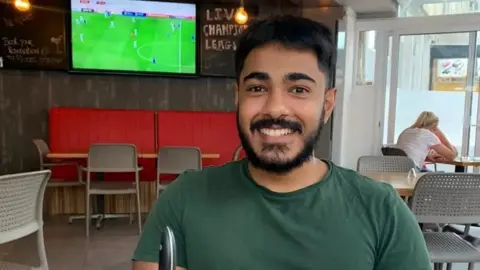 Family handout Anugrah Abraham, a man in his early 20s with black hair and thick stubble. He is wearing a green T-shirt and sitting in a bar with a football match on a TV in the background. He is smiling into the camera.