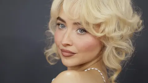 EPA Sabrina Carpenter poses with a neutral expression for a close-up shot in front of a black background. The silver strap of her dress can be seen on her shoulder, and her blonde hair is styled in a voluminous, curly style. 