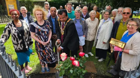 Ipswich Amnesty Group Ipswich Mayor Elango Elavalakan holds a spade, helping plants roses in a garden with a crowd gathered round.