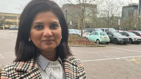 A picture of a woman with dark brown hair. She is wearing a white and black striped jacket with an orange stripe too. She is also wearing a white jumper underneath and is stood ina car park with buildings behind her.