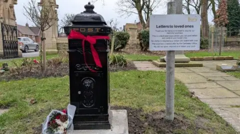 LDRS The black pillar postbox has a red ribbon tied around the top and a bouquet of flowers lies at the bottom on the right. Graves lies around it in the cemetery. A Bury Council sign on the right says "Letters to loved ones".