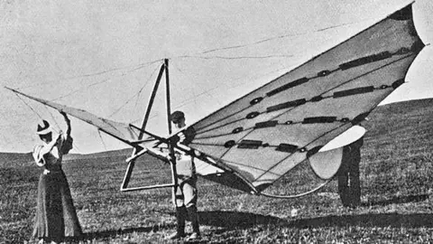 Philip Jarrett A black and white image of Percy and Ella Pilcher with the Bat Glider, a device with large wings. They are stood in a field holding the Bat Glider.