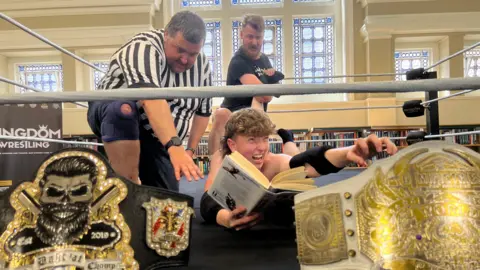 Jessica Bayley/BBC Image shows a boy lying on the floor of a wrestling ring reaching out with one arm and in his other hand he is holding a book. A referee is next to him and behind him another man is holding his leg in a wrestling hold. In the foreground there are wrestling title belts, a stained glass window is behind them and you can also see books on a bookshelf.