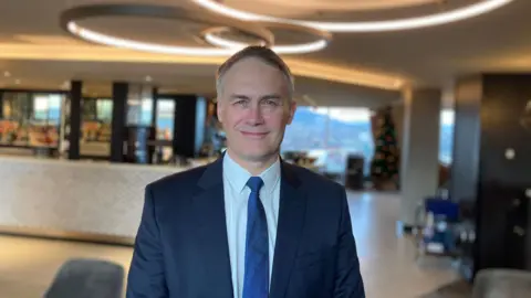 A man wearing a blue suit and blue tie smiles at the camera.
