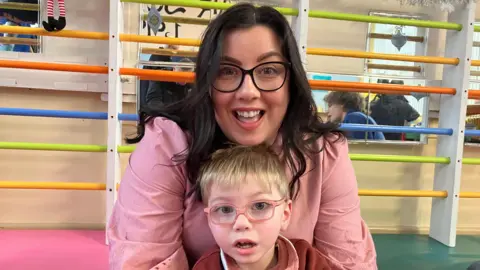 A woman with long dark hair is smiling at the camera. She is wearing dark rimmed glasses and a pink shirt. She has her arms around a young boy with blond hair and pink rimmed glasses.