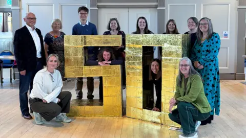 Wiltshire Community Foundation Ten woman and two men stood next to a giant gold 50 sign that comes up to about shoulder height. All are looking at the camera and smiling. They are inside a large hall with wooden floors and paneled walls.