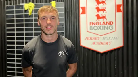 Tom smiles at the camera, his hands behind his back, an England boxing sign behind him. He has short blonde hair and is wearing a black T-shirt