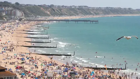 Andrew Matthews/PA Wire Bournemouth's beach with groynes stretching into the water. There are lots of people in the sea and packed onto the sand. It is a sunny day.