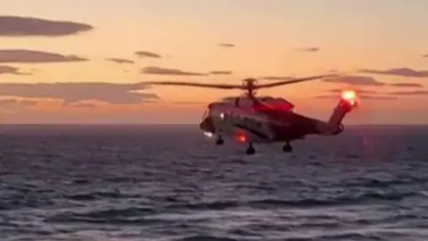The image shows a coastguard rescue helicopter hovering low over the sea close to a shoreline at dusk. In the foreground there are two parked vans beside a coastal road or car park, while waves are breaking near the shore below and a small figure can be seen in the water or surf; the helicopter's red lights are illuminated against a sunset sky, suggesting an ongoing maritime rescue or emergency response operation near a beach.