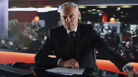 Actor Martin Clunes depicted as former BBC presenter Huw Edwards. Clunes is photographed in character wearing a black suit and silver watch. The actor is sat in a newsroom studio with pieces of paper in front of him and a newsroom with journalists working behind him.