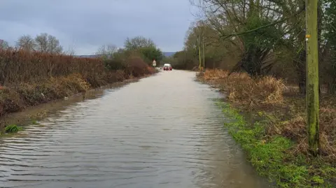 A long narrow stretch of flooded road lined with trees and hedgerows. In the distance, firefighters in orange waders are guiding a raft with a person on board away from a partly submerged white van.
