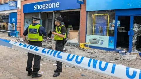 BBC Two police officers stand between a blue-and-white cordon and a badly damaged Shoezone shop in Hull city centre. Windows are broken and there is charred debris on the ground.
