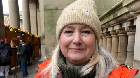 A woman with blonde hair wearing a beige hat. A green polo neck jumper and parka with orange lining can just be seen. She is standing outside a sandstone building with ornate stone pillars.