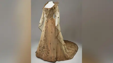 Royal Collection Enterprises Limited 2026 A gold dress hanging on a headless mannequin on a grey background.