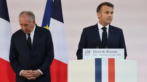 AFP via Getty Images Francois Bayrou knots his hands together and looks at the floor as he stands next to President Macron at an event in July.