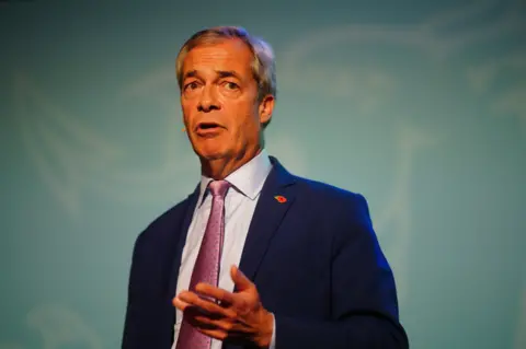 Nigel Farage wearing a blue suit, with a light blue shirt underneath and a pink tie. He is about to speak and has a mic on his face.