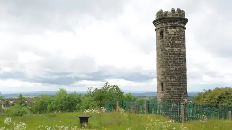 The tower stands on the right of the image with fencing around it and trees at a low level. A meadow-like field fills the remainder of the image with views spreading across the horizon.