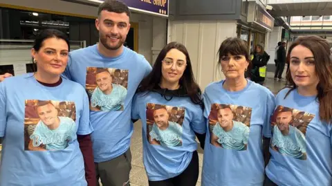Chloe Hilling, in the centre with dark hair and glasses, and members of her family in blue t-shirts with an image of her partner Gary on the front of the t-shirts. There are four women and one man, the man is second from the left.
