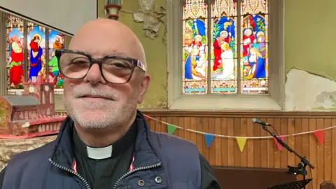 A middle-aged male vicar, with a bald head and light grey stubble, stands inside a church with a multi-coloured stained glass window in the background. He is wearing a blue gilet with his dog-collar and black smock visible. 