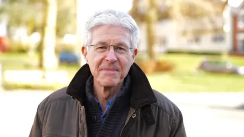 Simon Dedman/BBC A white-haired man standing outside. He is wearing glasses and smiling faintly. He is wearing a brown coat and a blue woollen cardigan and light blue shirt underneath. 