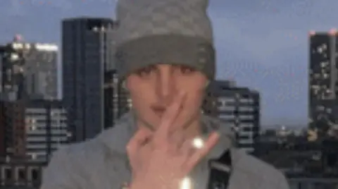 A teenage boy wearing a hoodie and a grey beanie stands on a balcony with skyscrapers in the background. He is holding his hand up in front of his face