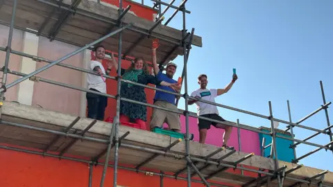 Four people - three men and one woman - stand on scaffolding on a very colourfully painted building, holding up paintbrushes and smiling against a clear blue sky.