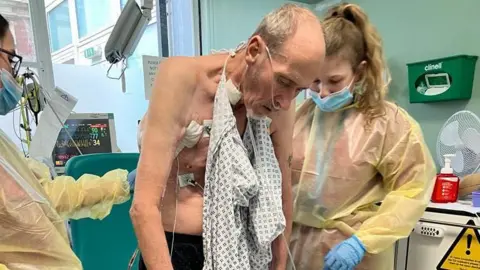 Paul Robinson is helped to stand up by nurses. He has a hospital gown hanging from his neck and has oxygen tubes in his nose.