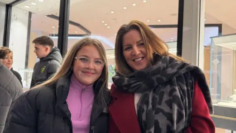 Lily-Rose and her mum Leanne standing side-by-side. Lily has long brown hair and is wearing a black coat with a purple sweater. Her mum also has long brown hair and is wearing a red coat and a patterned scarf.