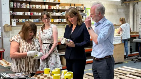 Chancellor Rachel Reeves, wearing a dark suit, smiles as she hold and examines a mug. She is standing in a ceramics workshop with two women and a man. A woman can be seen working in the background.