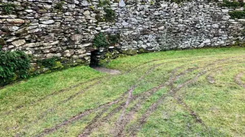 A curved stone wall is in the background. In the foreground is grass which has been torn up to mud by motorbike tyres. 