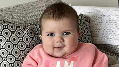 Mabli Hall sitting propped up in a grey sofa with black and grey patterned cushion. She is wearing a pink jumper.