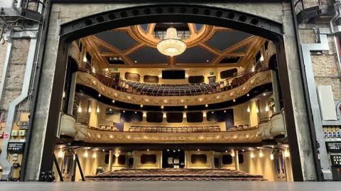 Jersey Opera House interior as seen from the stage. There are stalls on the lower tier, a middle circle and an upper circle of seats. The walls look yellow and gold in appearance from the use of lighting. There is a large chandelier hanging from the roof. 