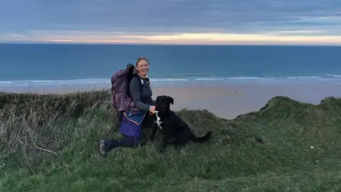 Handout Tara kneels on a grassy cliff overlooking a beach with a medium sized black dog kneeling by her side