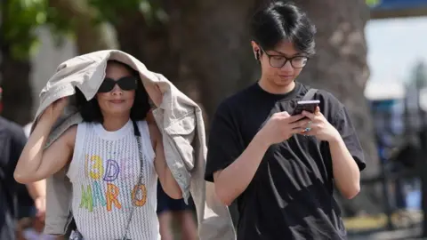 PA Media A woman holds a jacket over her head during sunny weather as a man walks next to her on his phone.