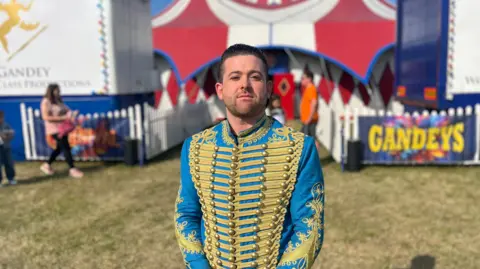 Joel Hatton, circus manager, faces the camera wearing a blue and gold circus jacket with buttons across the front. He looks serious but with a slightly wry smile. 