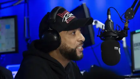 DJ Target in a black hat speaking into a Radio 1Xtra microphone in a blue studio with computers flanking him. 