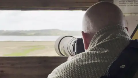Man with a big-long lens camera pointing at the lake in the distance.