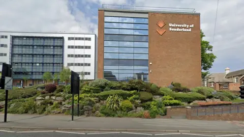 Google Brown brick building with windows reflecting blue sky. It has a large sign saying 'University of Sunderland' 