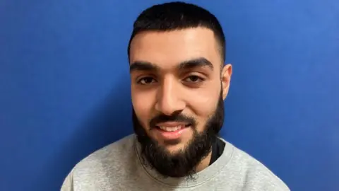 Alex Pope/BBC Fakhir, standing by a blue wall. He has a beard and is wearing a grey top. He has short dark hair.