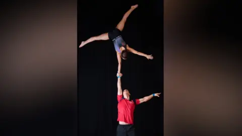 Jade Gibbs Photography A man and woman gymnast mid-routine. The woman is upside down, cartwheeling above the man, and he is supporting her by holding up his right arm. She 