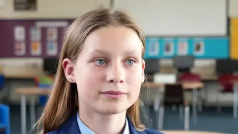 Shaun Whitmore/BBC A head and shoulders image of a young woman. She is wearing a navy blazer over a smart shirt. She is looking away from the camera. She is sitting in a classroom and there are chairs and tables behind her.