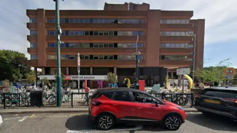 Google Five-storey brick-built building with Watford Junction on a white sign above the entrance. There are people on bicycles on the paved area in front of the station. There is a black railing separating the station from the road, on which a red car is stationary.