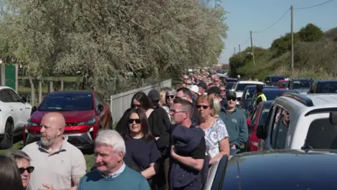 A queue of men and women of all ages goes back as far as the eye can see, along a lane with cars parked either side and a sea embankment on the right.
